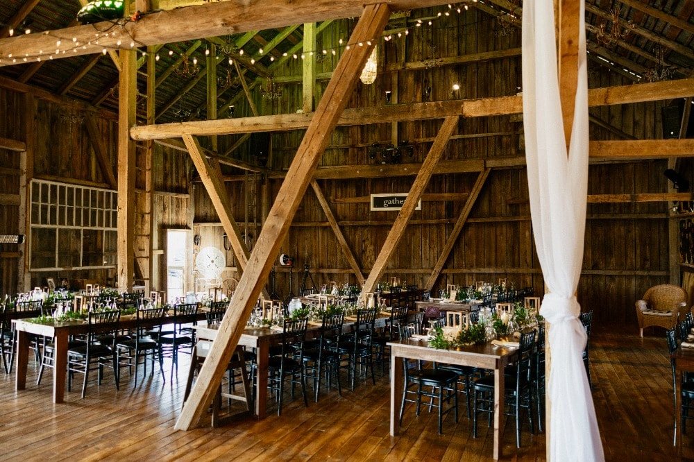 The main floor of the barn set for a seated dinner — farm tables and black chiavari chairs under hand-hewn trusses, chandeliers and string lights, with white drapery on the posts.